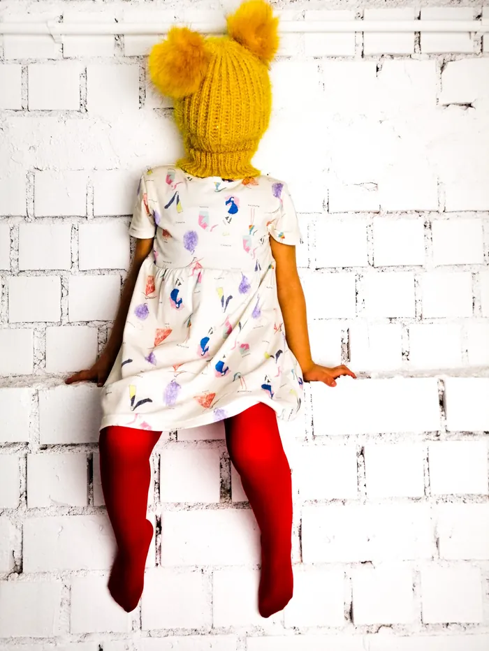 Niña sentada sobre un muro blanco con vestido estampado de dibujos, medias rojas y gorro de punto mostaza que le cubre la cara