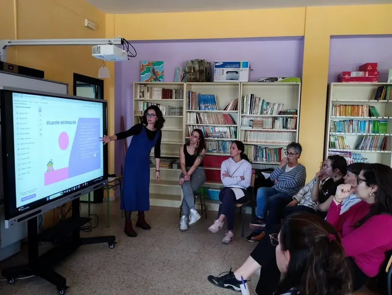 Pía Gutiérrez explica una presentación en una pantalla a un grupo de docentes sentado en la biblioteca de un colegio